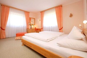 A hotel room with a wooden bed, two windows with curtains, a bedside table with a lamp, and a small stool. - Holiday apartment with 1 bedroom (Grainau)