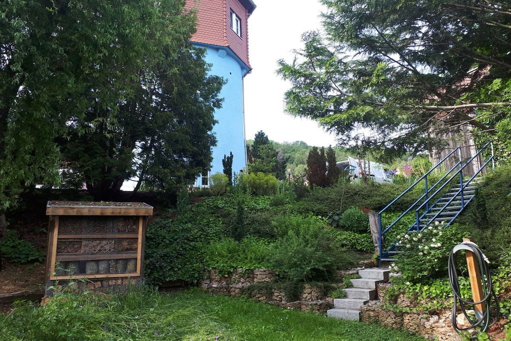 Ein Garten mit einer Steinsäule, einer Holzhütte und einer Treppe, die zu einem Gebäude mit rotem Dach führt.