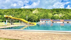 Piscina com escorregador amarelo, rodeada por um deck de tijolos, painéis solares em edifícios próximos e uma colina arborizada ao fundo.
