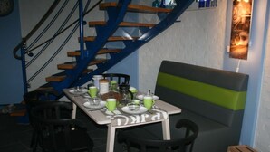Uma área de refeições com mesa posta para quatro pessoas, cadeiras, um banco e escada com degraus de madeira e corrimão azul.