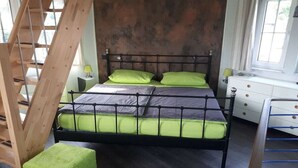 Cama com estrutura metálica e roupa de cama verde, escada de madeira, mesa de cabeceira branca com abajur e janela com cortinas.