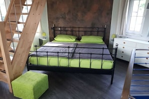 Een bed met metalen frame en groene beddengoed, een houten trap, een witte nachtkastje met een lamp, en een raam met gordijnen.