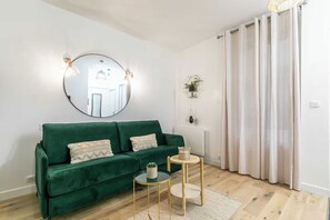 A living room with a green sofa, a round mirror, two small tables, and a view of a kitchen through a glass door. - Charming studio - 2P - Voltaire (Paris)