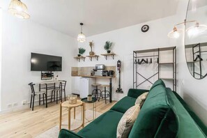 A modern living room with a green sofa, a flat-screen TV, a kitchen area with a coffee machine, and a dining setup. - Charming studio - 2P - Voltaire (Paris)