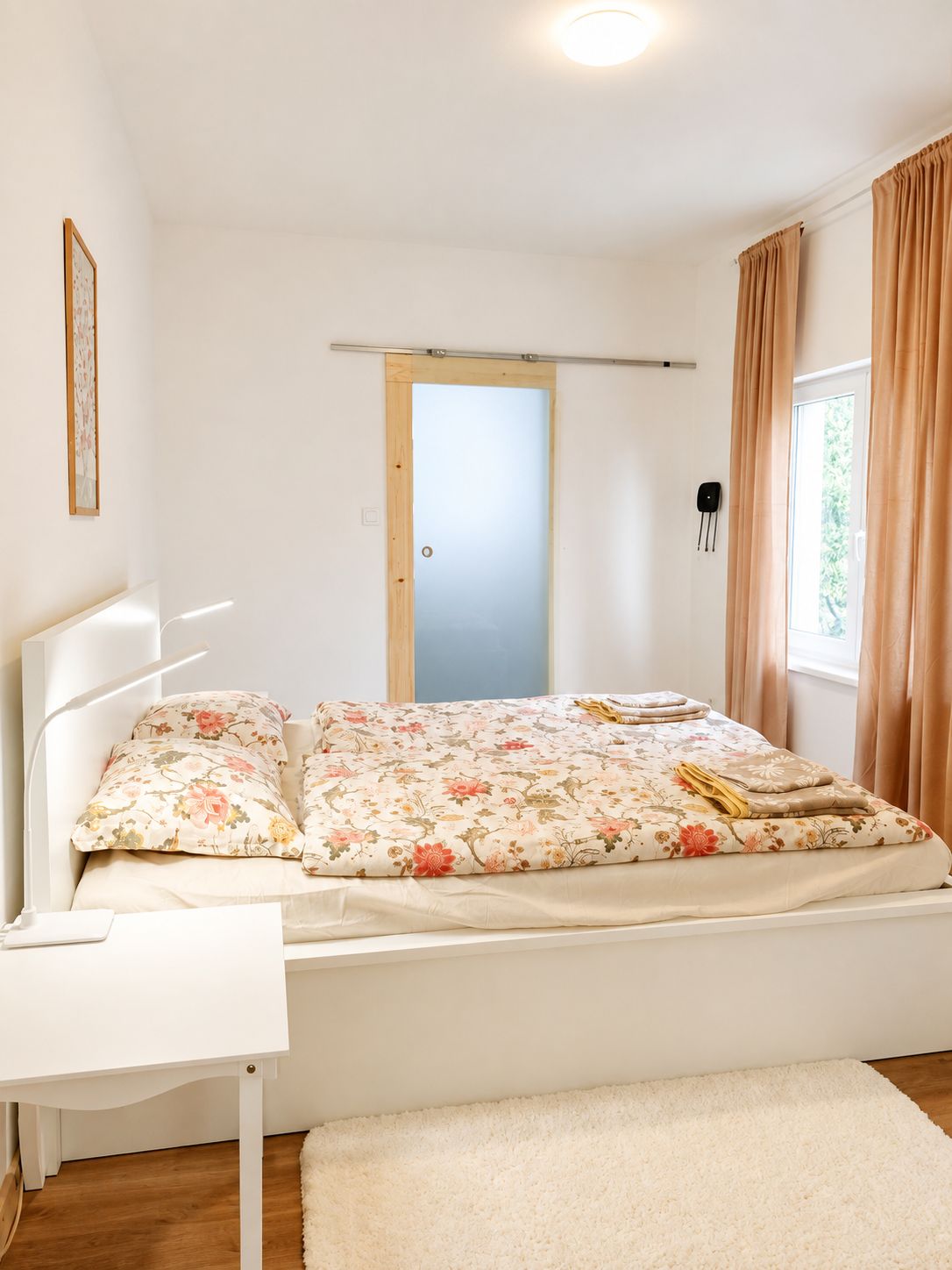 A bedroom with a bed, a bedside table, a window with curtains, a door, and a wall-mounted light fixture.
