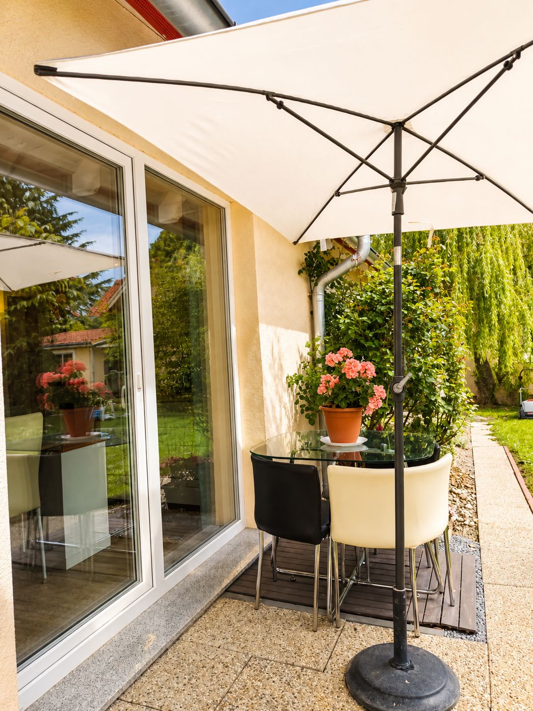 Eine Terrasse im Freien mit einem Glastisch, zwei Stühlen, einem Topfpflanzen und einem großen Sonnenschirm.