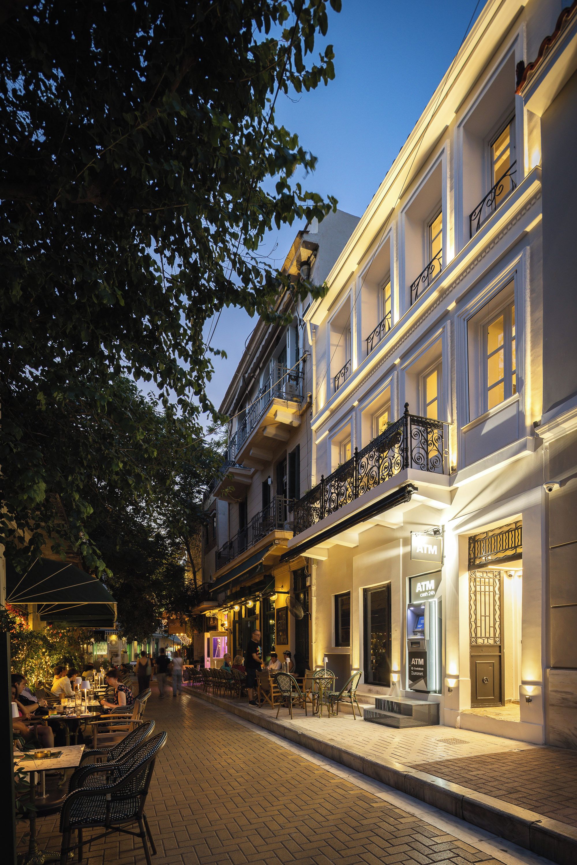 A street with outdoor seating and a building with an ATM on the ground floor.