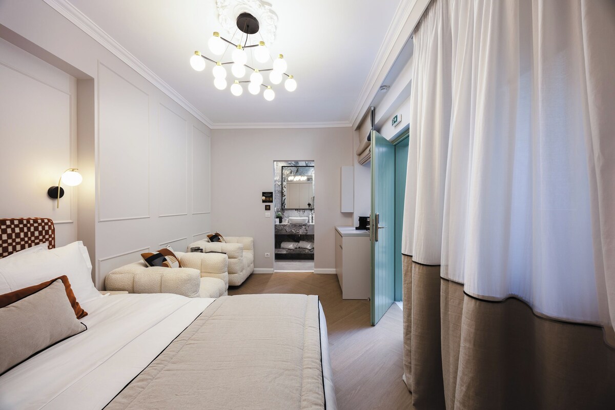 A hotel room with a large bed, a seating area with ottomans, a bathroom with a mirror, and a kitchenette.