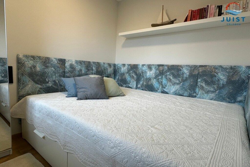 A bedroom with a bed, pillows, a headboard with a leaf pattern, a shelf with books, and a small decorative item.