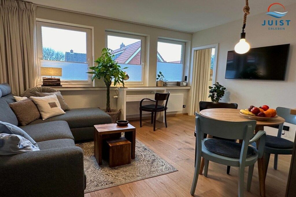 Ein modernes Wohnzimmer mit einer Couch, einem Esstisch mit Stühlen, einem an der Wand montierten Fernseher und großen Fenstern mit Vorhängen.