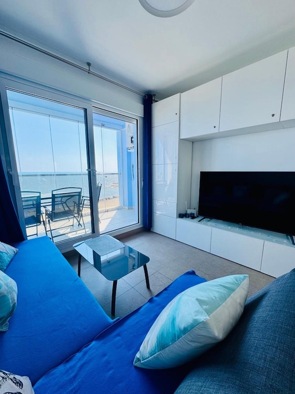 Una sala de estar moderna con un sofá, una mesa de centro y un televisor de pantalla plana. Hay una puerta corrediza de vidrio que da a un área exterior con vista al mar.