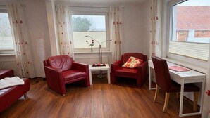 Une pièce avec un plancher en bois, deux fauteuils en cuir rouge, une petite table blanche avec une lampe, un bureau avec une chaise et une fenêtre avec des rideaux.