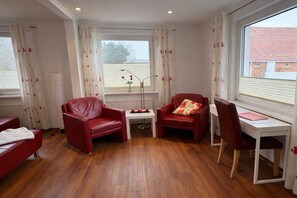 A room with a wooden floor, two red leather chairs, a small white table with a lamp, a desk with a chair, and a window with curtains. - Holiday apartment with 3 bedrooms (Norderney)