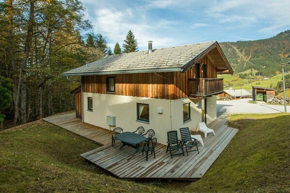 A wooden cabin with a deck, outdoor seating, and a view of the surrounding forest and hills.