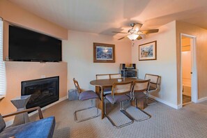 A living room with a flat-screen TV, fireplace, dining table with chairs, ceiling fan, and two framed pictures on the wall.