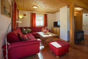 A cozy living room with a red sofa, a wooden ceiling, a television, and a wood-burning stove. - Cabin in Annaberg with swimming pool (Annaberg)