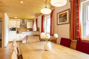 A wooden dining table with a vase of flowers, a kitchen area with white cabinets, and a window with red curtains. - Cabin in Annaberg with swimming pool (Annaberg)