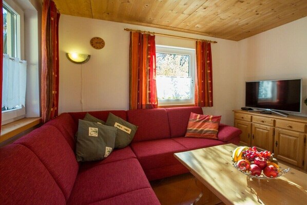 Un salon avec un canapé rouge, une table en bois sur laquelle se trouvent des fruits, un téléviseur et une fenêtre avec des rideaux.