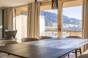 A dining area with a large table and chairs, large windows with curtains, and a view of a mountainous landscape. - Premium penthouse with 3 bedrooms (Oberndorf in Tirol)