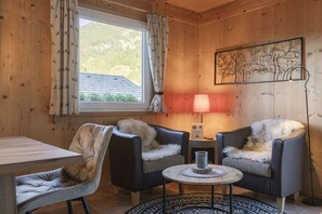 Ein gemütlicher Raum mit Holzwänden, einem Esstisch, zwei Sesseln mit Felldecken, einem kleinen runden Tisch mit Kerze und einem Baumgemälde an der Wand.