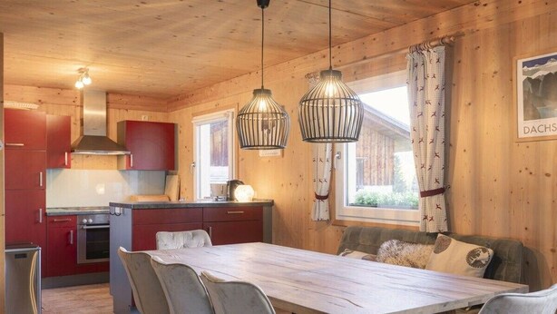 Interior de una cabaña de madera con comedor, cocina y una ventana con cortinas.
