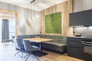 Una cuina moderna amb forn integrat, zona de menjador amb taula de fusta i cadires grises, i un element decoratiu a la paret de color verd.