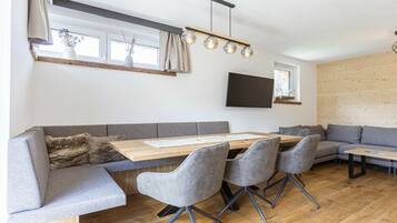 A modern living space with a wooden dining table, grey upholstered seating, and a flat-screen TV mounted on the wall.