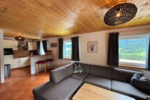 Een gezellig chaletinterieur met een houten plafond, een bank, een eettafel en een keukenhoek.