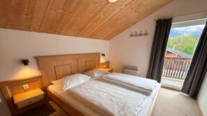 A bedroom with a wooden ceiling, a bed with white bedding, a nightstand, a wall-mounted heater, and a view of a balcony with a wooden railing. - Cabin in Annaberg with swimming pool (Annaberg)