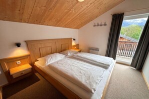 A bedroom with a wooden ceiling, a bed with white bedding, a nightstand, a wall-mounted heater, and a view of a balcony with a wooden railing. - Cabin in Annaberg with swimming pool (Annaberg)