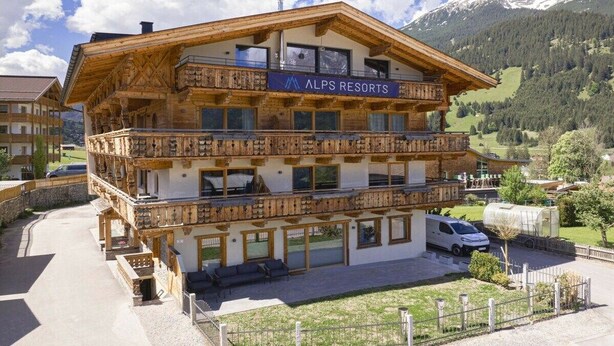 Ein mehrstöckiges Holzgebäude mit Balkonen, beschriftet mit 'ALPS RESORTS', inmitten einer gebirgigen Landschaft.