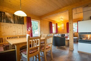 A wooden cabin interior with a dining area, sofa, fireplace, and large windows.