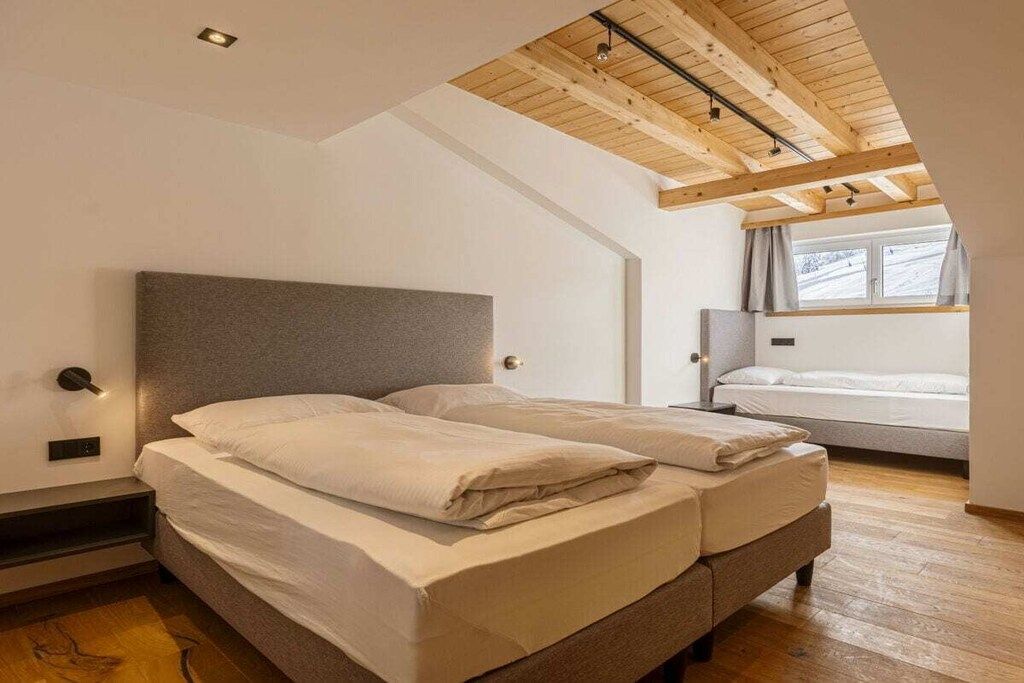 A bedroom with a large bed, a smaller bed, wooden ceiling beams, and a window with curtains.