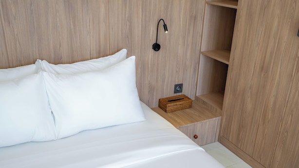 Una cama bien hecha con sábanas y almohadas blancas, una lámpara de noche, una mesita de noche de madera y una cesta de mimbre.