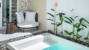 Un área de estar al aire libre y moderna, con un sofá blanco y cojines, una pequeña mesa de madera y una jardinera rectangular con plantas verdes y una sola flor.