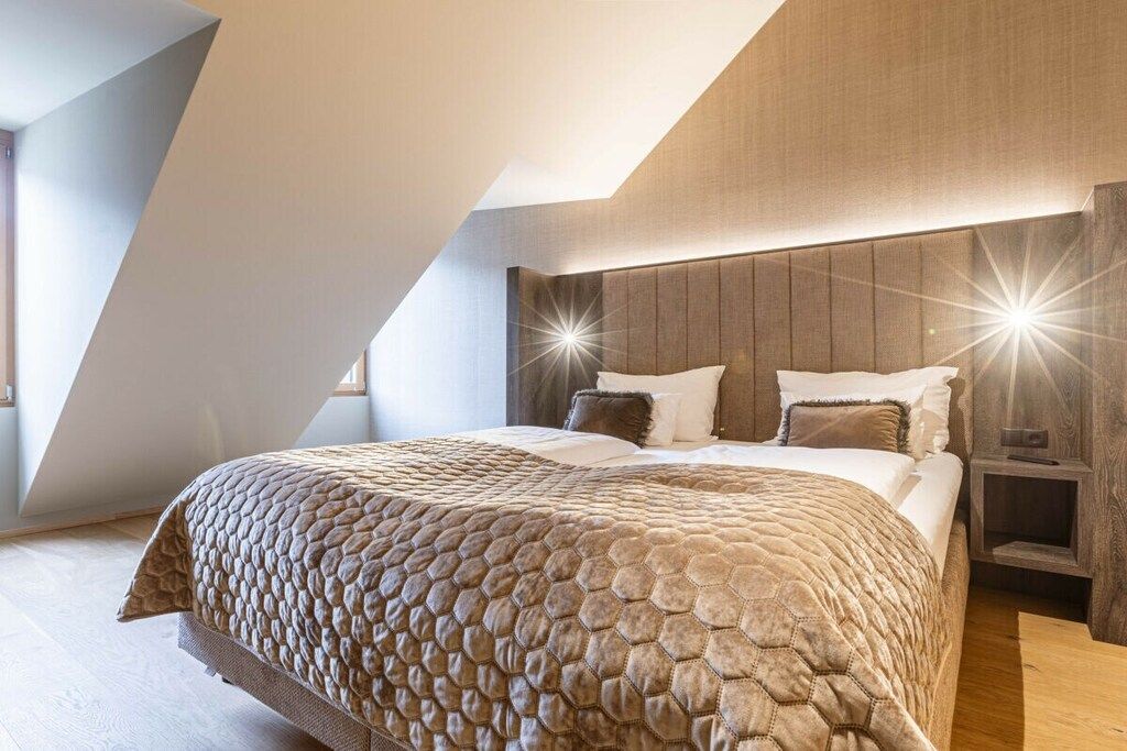 Une chambre avec un plafond en pente, un grand lit recouvert d’une couverture à motifs, et une table de chevet avec une lampe.