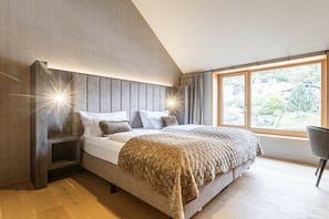 A modern bedroom with a large bed, a nightstand, a chair, and a window with a view of greenery. - Apartment in Schruns with swimming pool (Schruns)