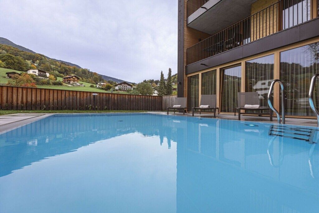 Une piscine avec une terrasse en bois, un bâtiment moderne doté de grandes fenêtres et une vue sur les maisons au loin.
