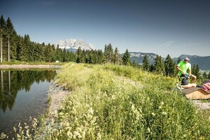 Eine Person sitzt auf einer Matte am See, im Hintergrund eine Berglandschaft.