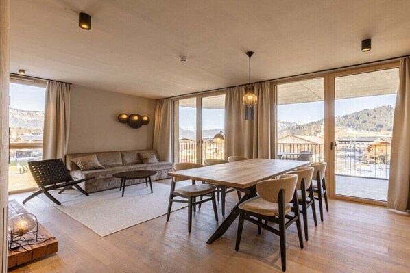Ein geräumiges Zimmer mit einem Holzespeisetisch, Stühlen, einer Couch und einem großen Fenster mit Blick auf eine Berglandschaft.