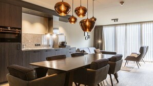 Une cuisine moderne avec un coin repas, comprenant une grande table et des chaises, un éclairage suspendu et une vue sur l’extérieur grâce à de grandes fenêtres.