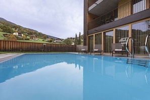 A swimming pool with a wooden deck, a modern building with large windows, and a view of houses in the background. - Apartment in Schruns with swimming pool (Schruns)
