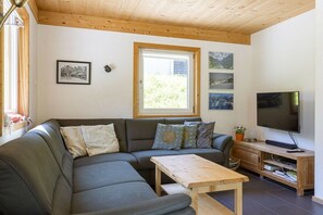 Un salon moderne avec un canapé d’angle, une table basse en bois, un téléviseur à écran plat et une fenêtre donnant sur un paysage verdoyant.