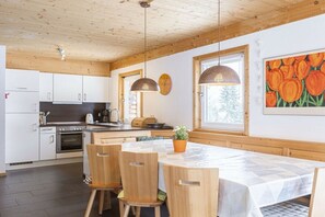 A modern kitchen with wooden cabinets, a dining table with chairs, and a large window with a view of trees. - Cabin in Annaberg with swimming pool (Annaberg)