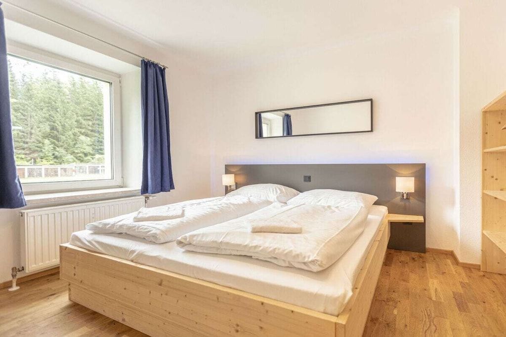 Ein Zimmer mit einem hölzernen Bett, einem fenster mit Vorhängen, einem wandmontierten Spiegel und einem Nachttisch mit einer Lampe.