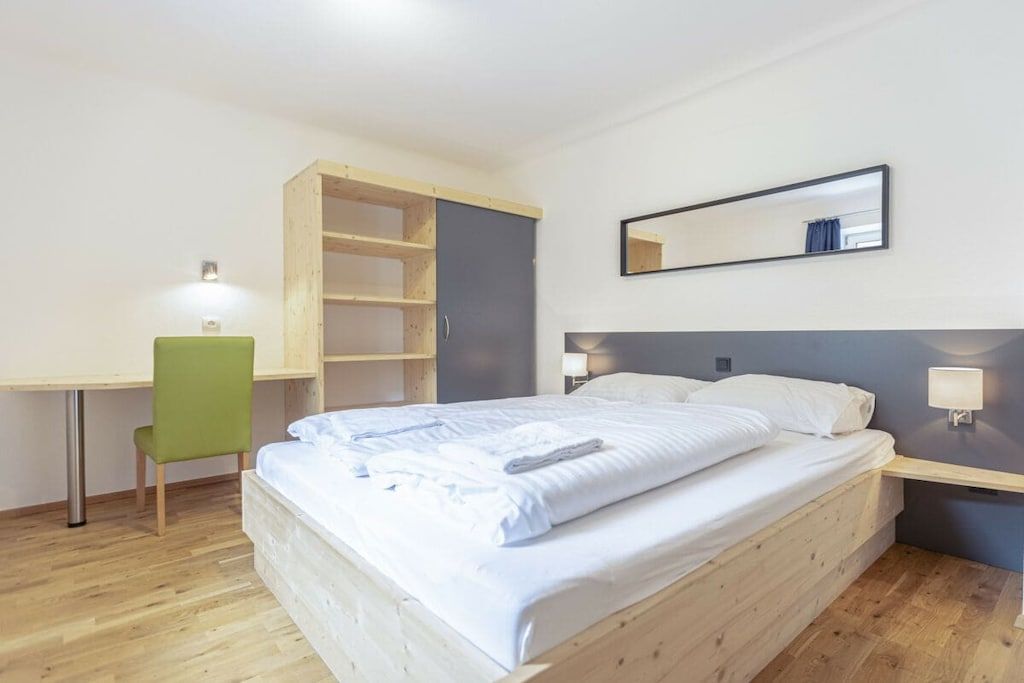 A single-bed room with a wooden headboard, a desk with a chair, a bedside table with a lamp, and a mirror on the wall.