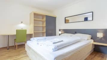 A single-bed room with a wooden headboard, a desk with a chair, a bedside table with a lamp, and a mirror on the wall.