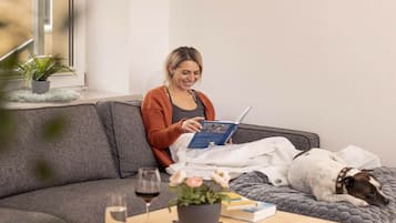A person sitting on a grey sofa with a book, a dog lying beside them, and a table with a glass of wine and a potted plant.