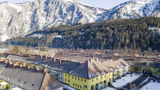 Un borgo con architettura tradizionale, un edificio giallo centrale con archi e montagne innevate sullo sfondo.