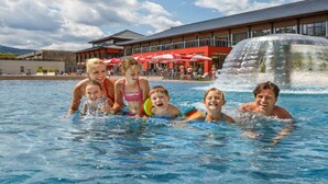 Una piscina con persone a nuotare, un edificio sullo sfondo con ombrelloni rossi e un elemento d'acqua a destra.